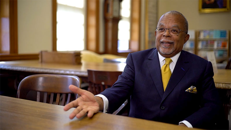Henry Louis Gates, Jr sitting at a table in suit and yellow tie discussing the Finding Your Roots - The Seedlings project